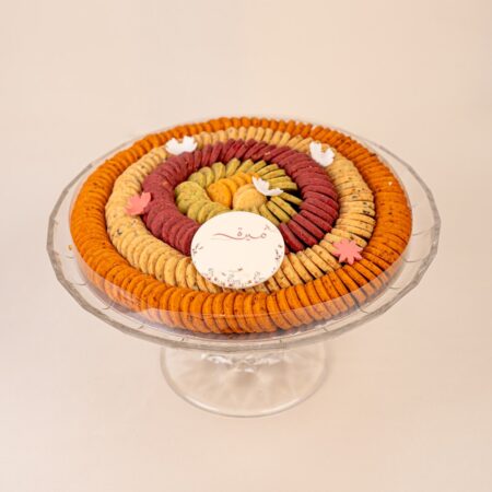 Round cookie tray with orange, red and green cookie rings under a clear lid on a crystal cake stand