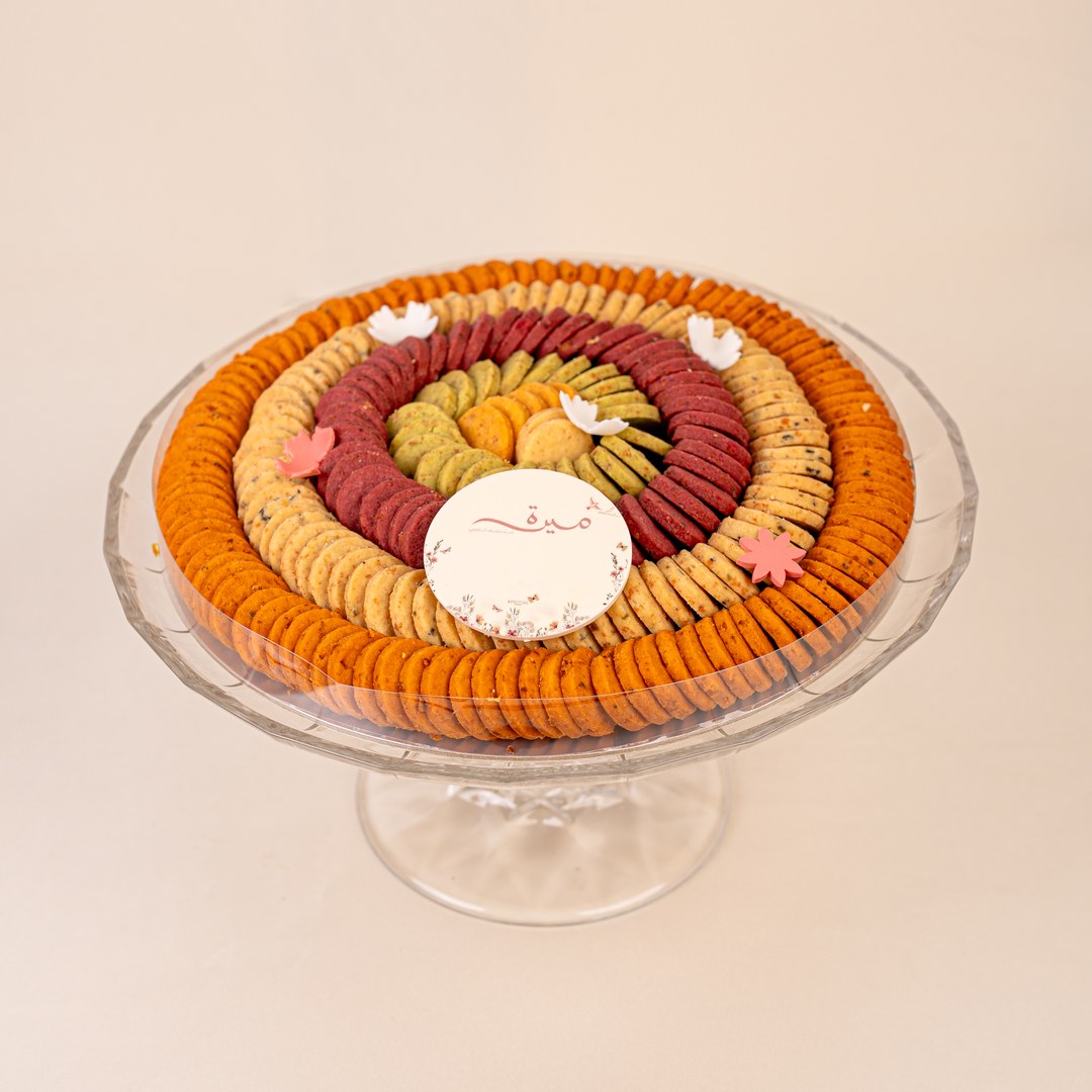 Round cookie tray with orange, red and green cookie rings under a clear lid on a crystal cake stand