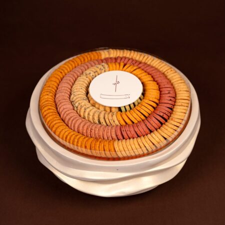 Luxury cookies tray with rings of orange, pink, golden and seeded cream cookies under a clear lid on a cream wave stand